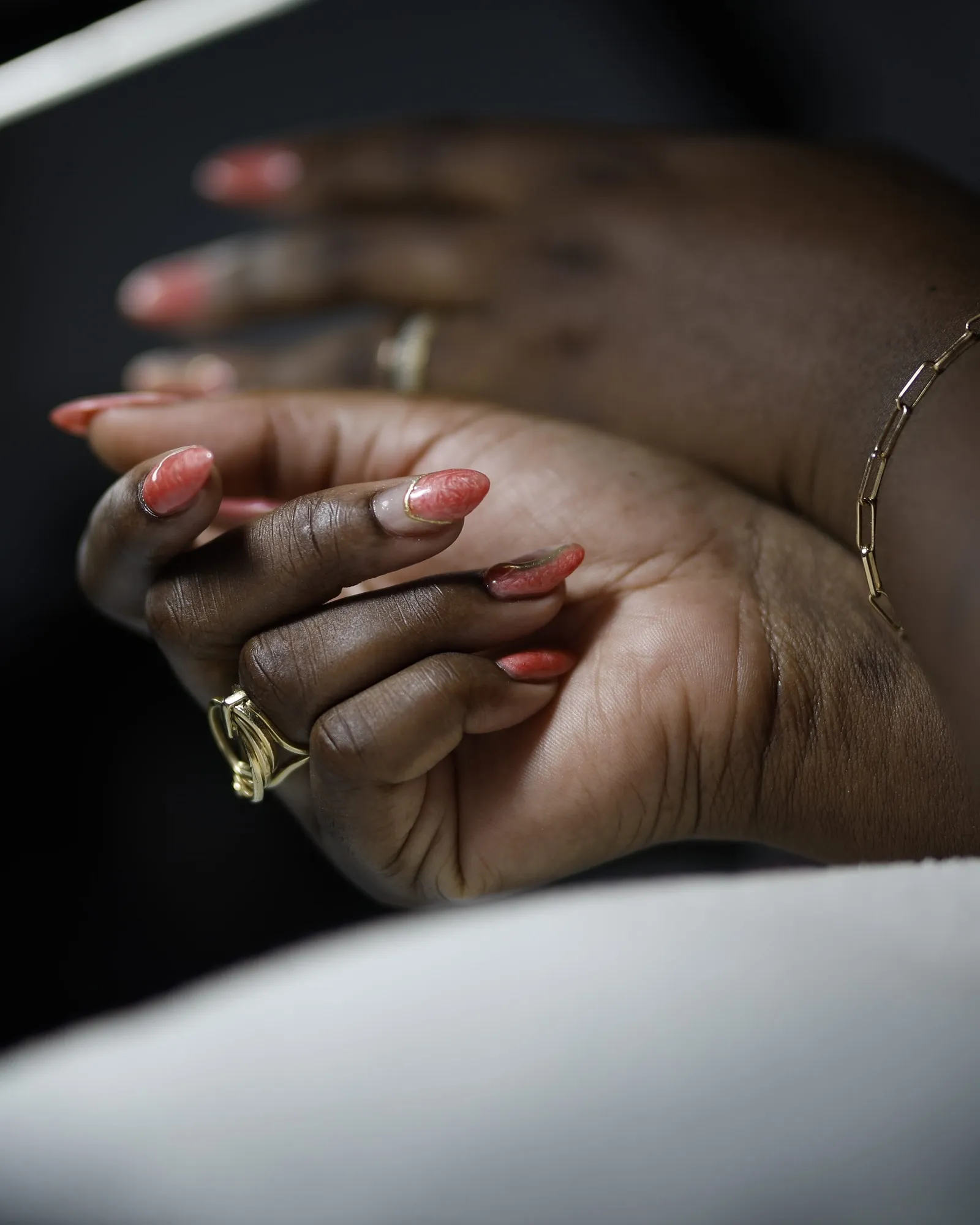 Deux mains aux ongles corail en amande, bague et bracelet dorés, posées sur un linge blanc