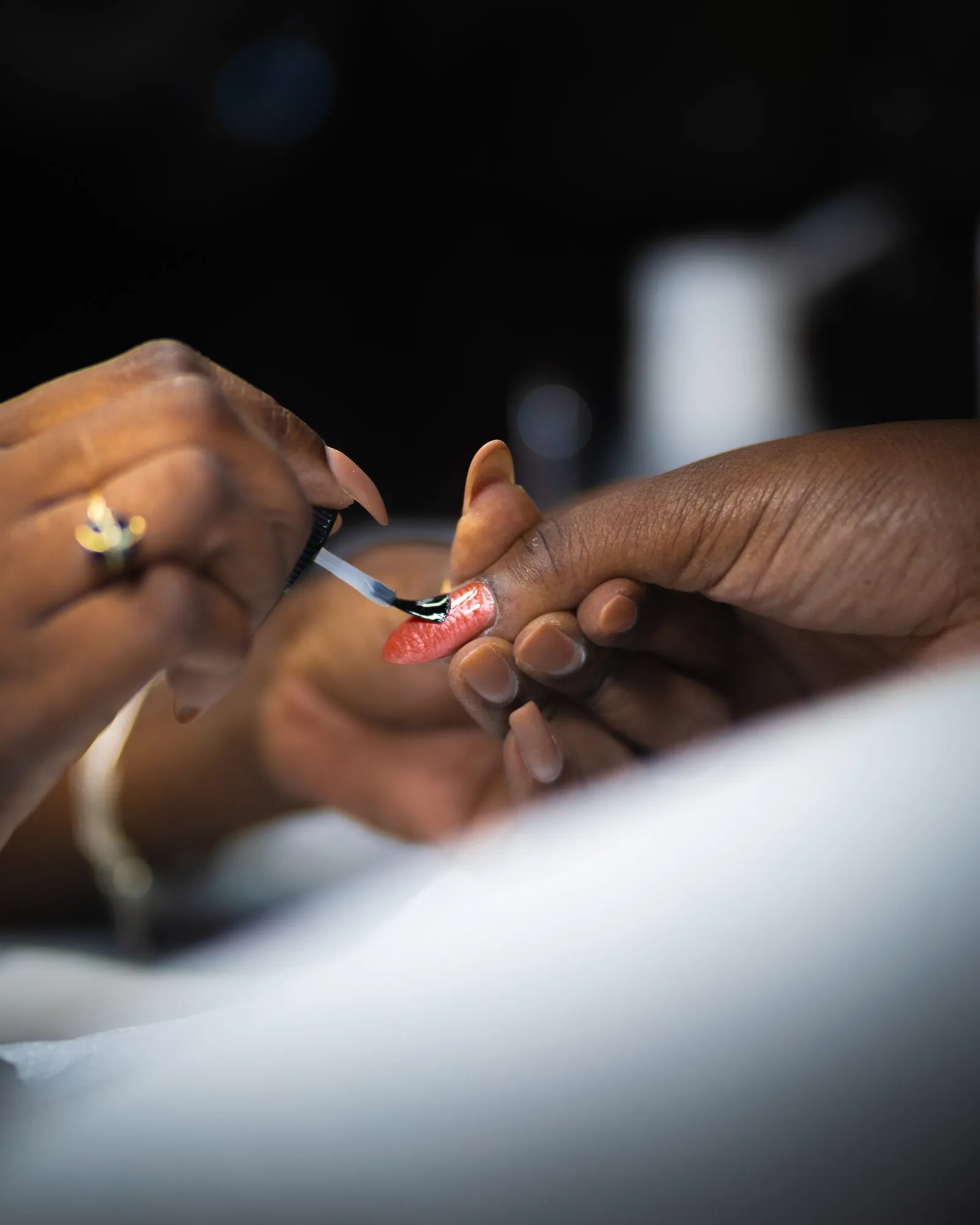 Une main baguée d'or applique au pinceau un gel corail sur l'ongle d'une cliente, au-dessus d'un linge blanc