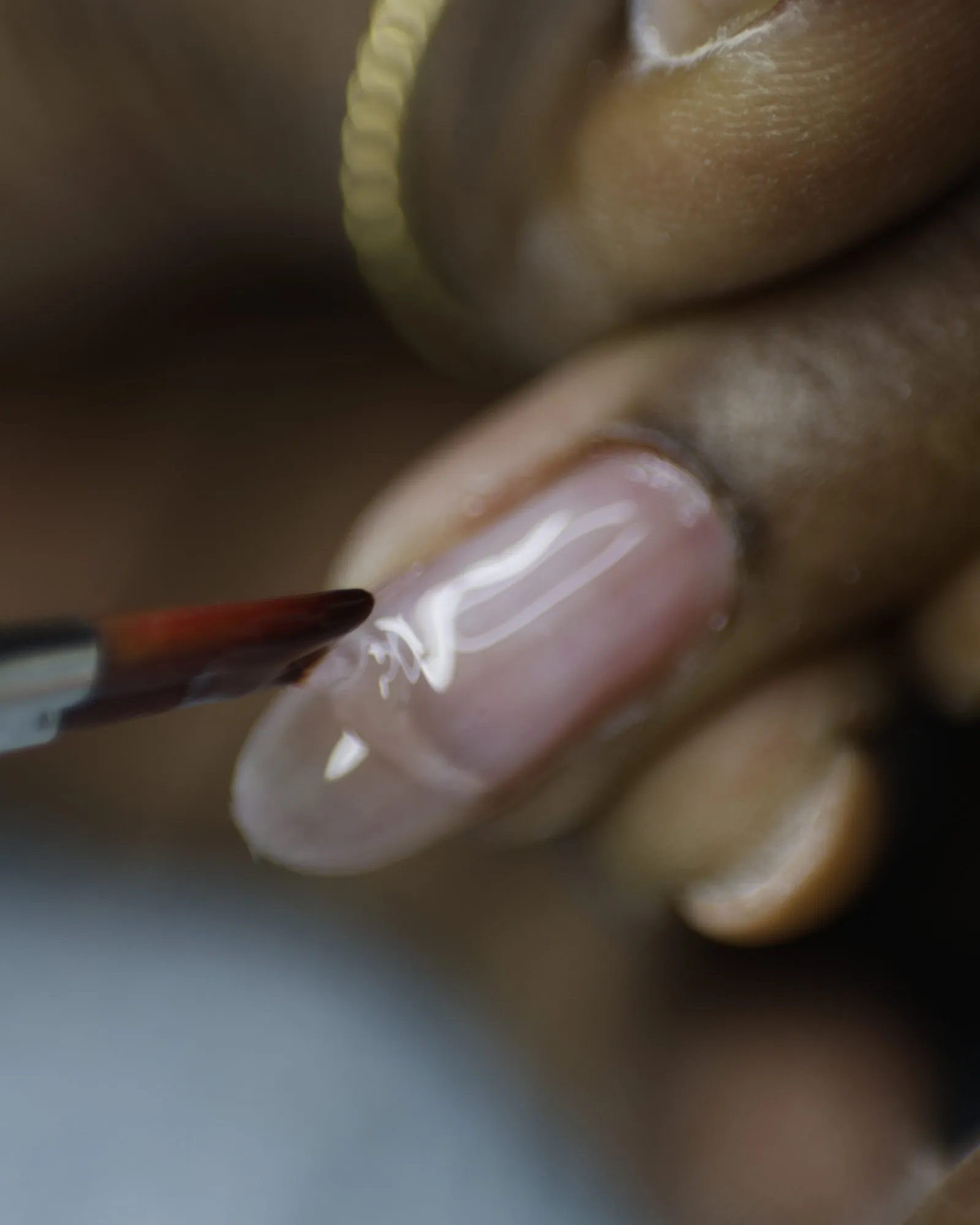 Gros plan d'un pinceau déposant le gel sur un ongle naturel, main baguée d'or en arrière-plan